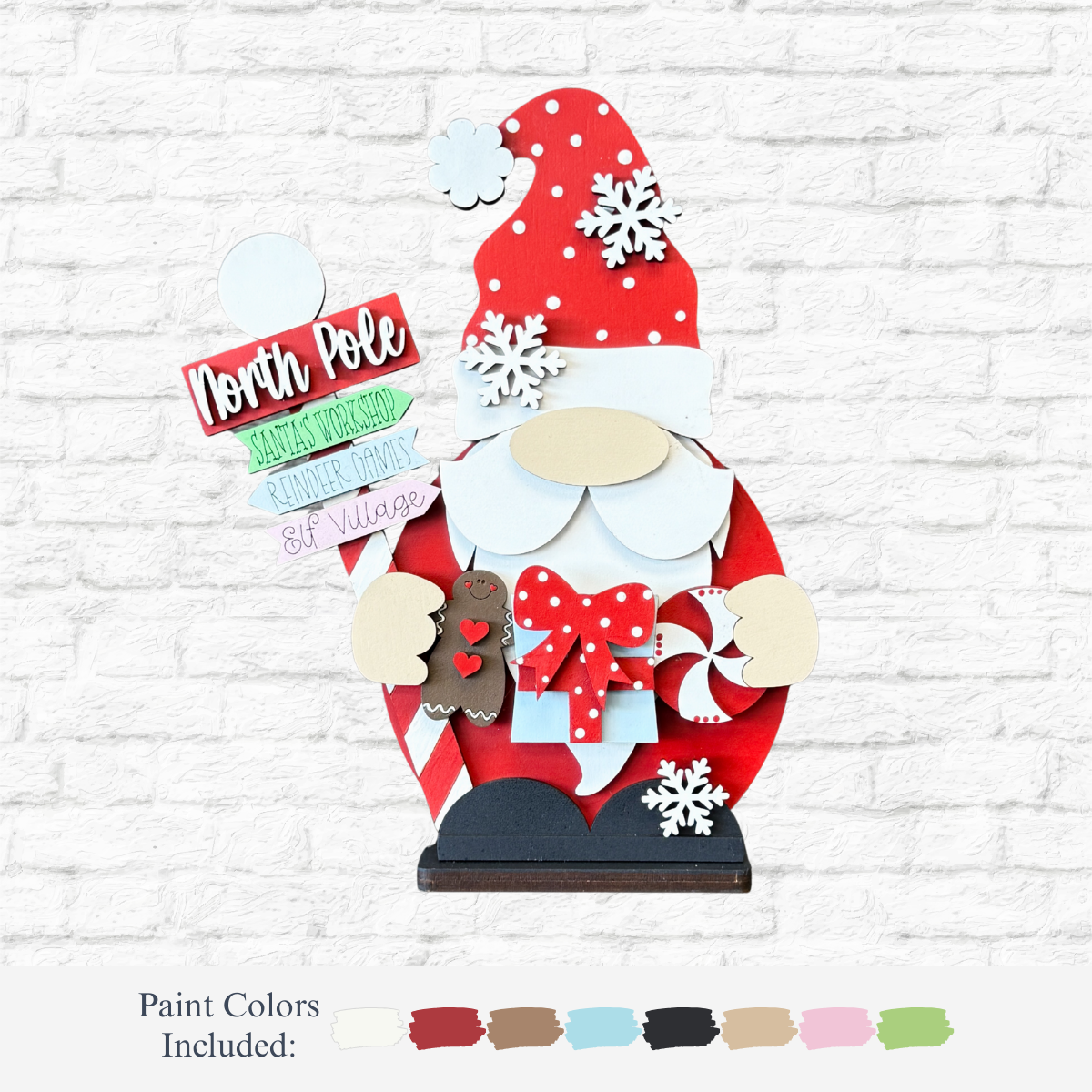 The Rustic Rhode's DIY North Pole Santa Gnome features a red-hatted gnome with holiday signs, gifts, treats, and snowflakes—ideal for festive home décor. Shown with red, brown, beige, green, blue, white & pink on a white brick background.