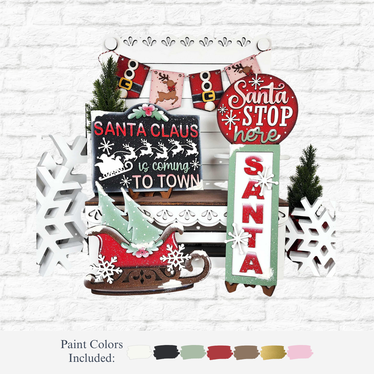 The Rustic Rhode’s DIY North Pole Tiered Tray Set features festive wooden decorations—signs, a sleigh, snowflakes, and pine trees—arranged on a white bench with paint color samples displayed below against a white brick wall.