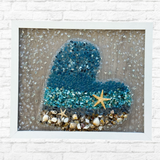 The Rustic Rhode DIY Ocean Heart Sea Glass Frame features blue and clear glass, shells, and starfish on a textured background, adding ocean-inspired charm to your space. Perfect coastal decor for any room.