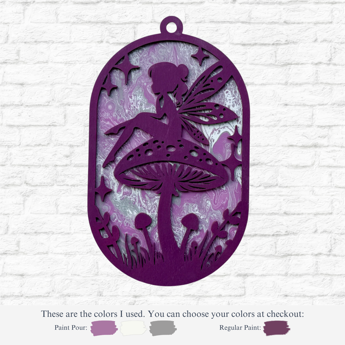 The Rustic Rhode’s DIY Paint Pour- Fairy is an oval purple ornament with a fairy on a mushroom, set against a marbled purple and silver background. Choose your paint colors for a personalized art experience or create your own custom kit!.