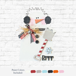 The Rustic Rhode’s DIY Prim Snowman kit features a laser cut snowman with a carrot nose, black earmuffs, plaid scarf, straw bow, striped shovel, and “Brrr” with snowflake accents—paint swatches included for your next winter craft!.