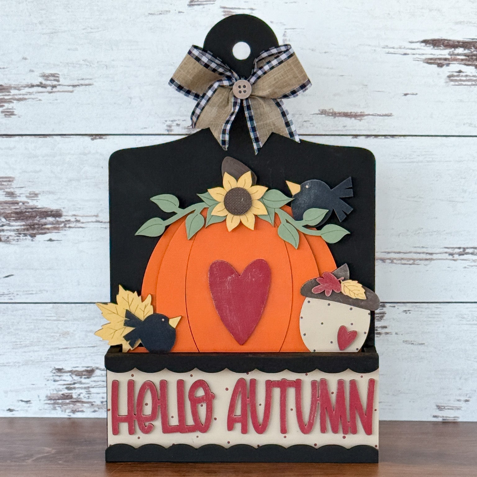 The Rustic Rhode’s DIY Pumpkin Season 2 Piece Insert features a pumpkin with a red heart, sunflowers, leaves, and blackbirds. This Handmade Craft Kit includes a plaid bow and HELLO AUTUMN for your Interchangeable Window Frame.
