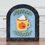 Decorative wall art with a pumpkin-themed mug and whipped cream on a wooden surface.