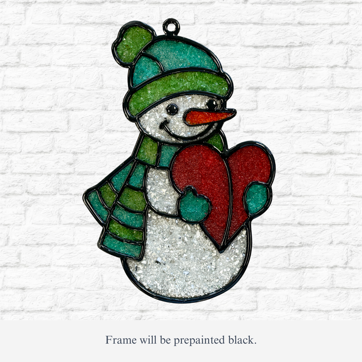 The Rustic Rhode DIY Snowman Faux Suncatcher features a smiling snowman with a green hat and scarf, holding a red heart. The prepainted black wood frame creates a charming resin suncatcher effect against any backdrop.
