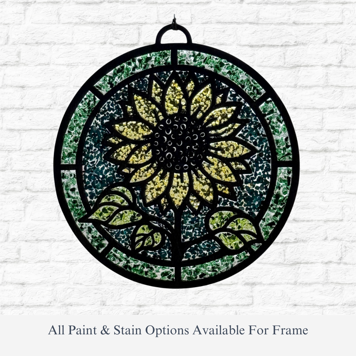 The Rustic Rhode DIY Sunflower Faux Suncatcher—a round panel with a bright yellow sunflower, green leaves, and black frame—hangs on a white brick wall. Text reads: “All Paint & Stain Options Available For Frame.”.