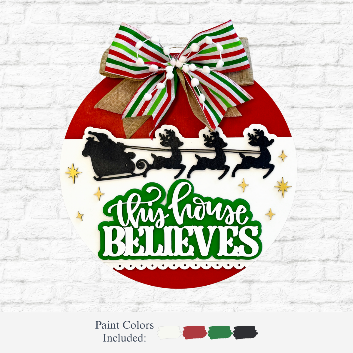 The Rustic Rhode’s DIY This Home Believes Door Hanger is a round, laser-cut wood sign in red, white, and green with a ribbon bow, Santa’s sleigh and reindeer silhouette, plus bold green text—ideal as a festive holiday decoration or craft kit.