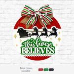 The Rustic Rhode’s DIY This Home Believes Door Hanger is a round, laser-cut wood sign in red, white, and green with a ribbon bow, Santa’s sleigh and reindeer silhouette, plus bold green text—ideal as a festive holiday decoration or craft kit.