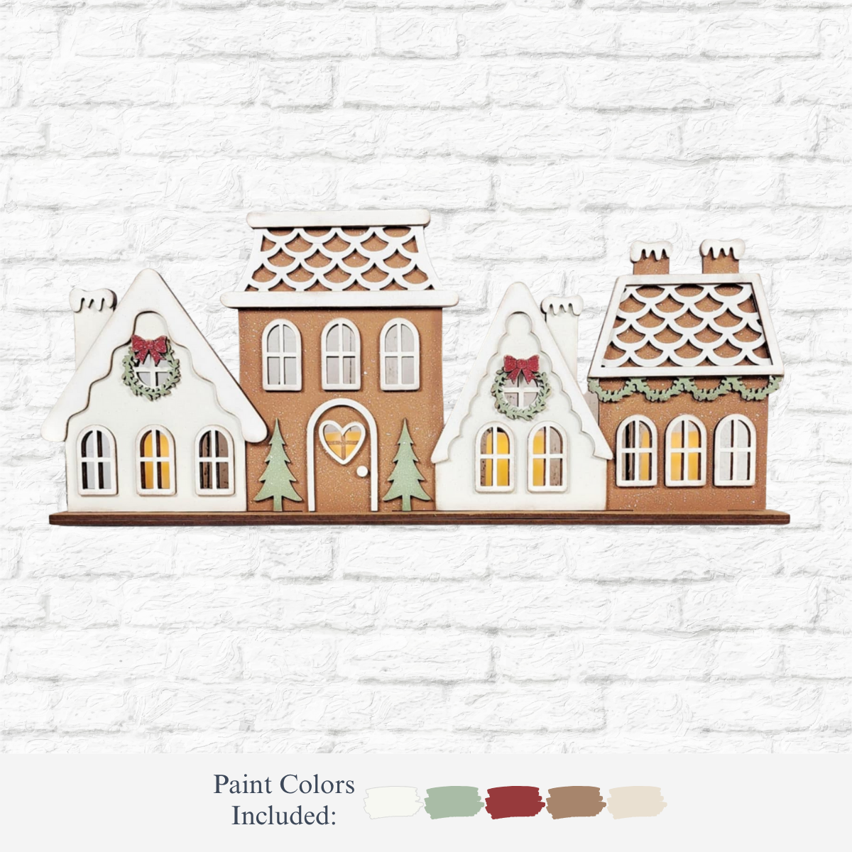 The Rustic Rhode’s DIY Victorian Gingerbread Village Shelf Sitter features three festive houses with wreaths, pine trees, and glowing windows. Four paint colors—cream, green, red, and brown—complete this charming holiday craft kit.