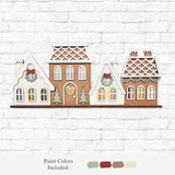 The Rustic Rhode’s DIY Victorian Gingerbread Village Shelf Sitter features three festive houses with wreaths, pine trees, and glowing windows. Four paint colors—cream, green, red, and brown—complete this charming holiday craft kit.