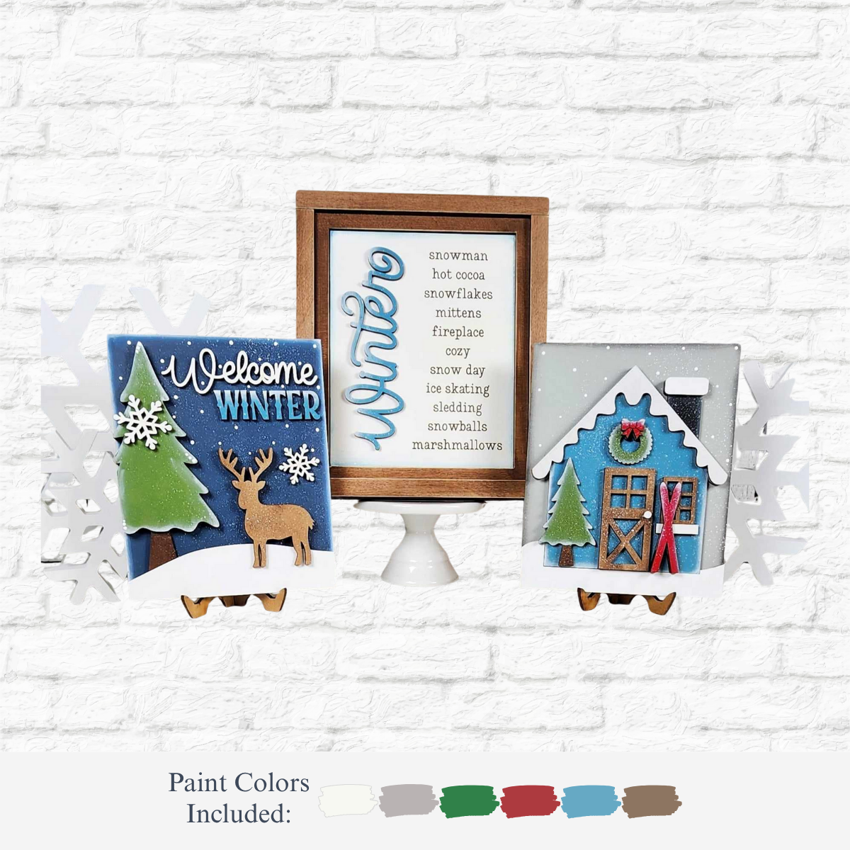 The Rustic Rhode’s DIY Welcome Winter Sign Trio features three home décor crafts on stands: a tree and reindeer sign, a framed winter words print, and a snowy blue house. Five paint colors—white, brown, blue, green, and red—are included below.