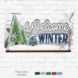 The Rustic Rhode DIY Welcome Winter Word Art features a green pine tree, small house, snowflakes, and "Welcome Winter" in white and blue on a white brick background—perfect for personalized home decor projects. Paint colors shown below.