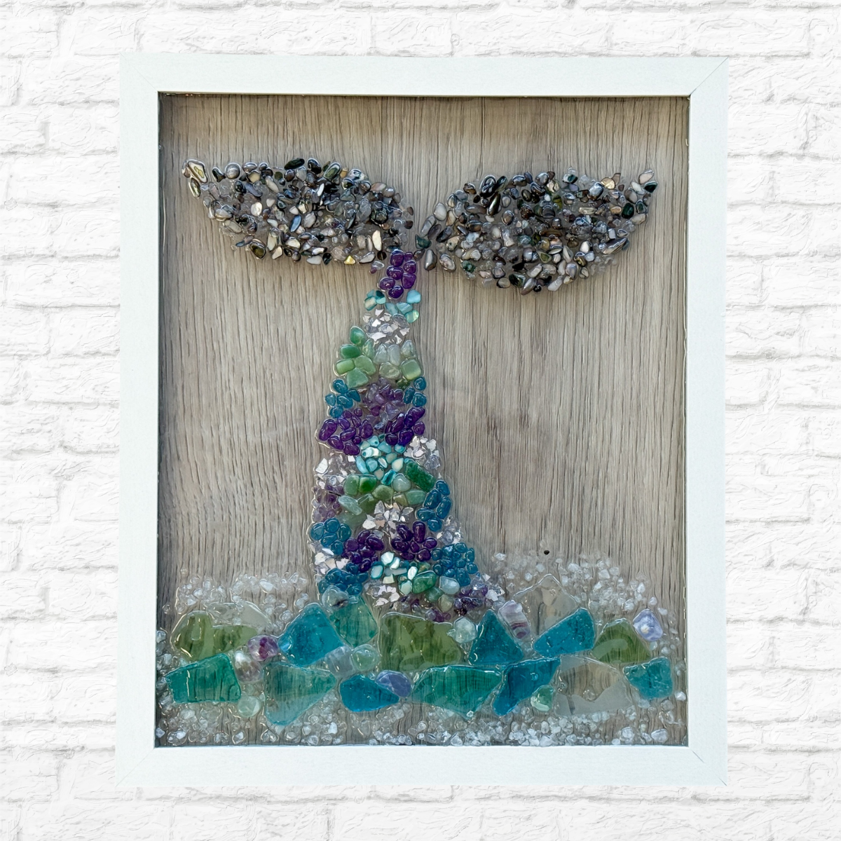The Rustic Rhode’s DIY Whales Tail Sea Glass Frame features a whale tail made from colorful sea glass and stones on wood, with blue and green glass waves, displayed against a white brick wall—ideal for ocean lovers or DIY inspiration.