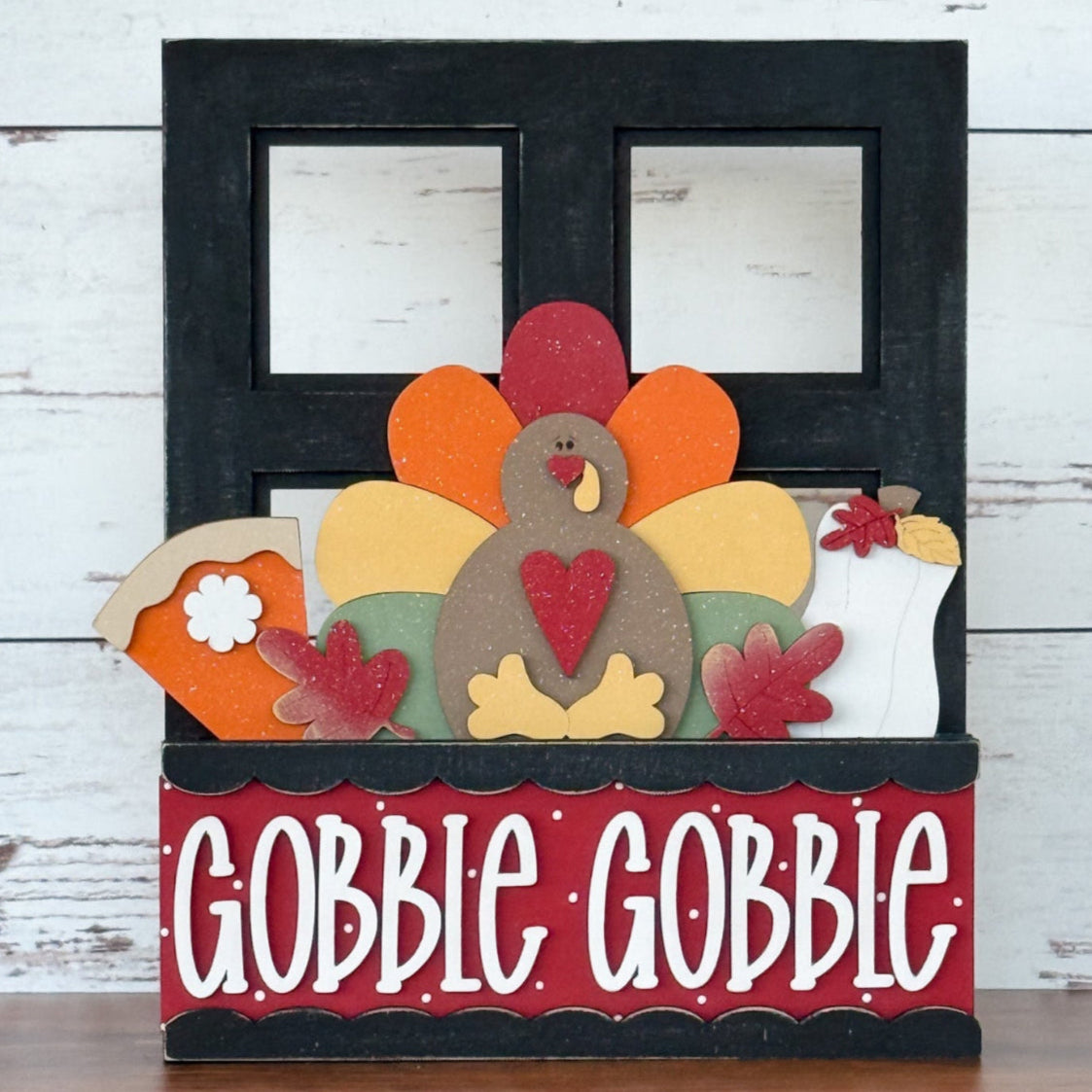 The Rustic Rhode DIY Interchangeable Window Frame showcases a colorful turkey, pie slice, and vase with “GOBBLE GOBBLE” in bold white letters on a red and black backdrop, all set against a rustic wood background.