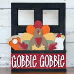 The Rustic Rhode DIY Interchangeable Window Frame showcases a colorful turkey, pie slice, and vase with “GOBBLE GOBBLE” in bold white letters on a red and black backdrop, all set against a rustic wood background.