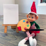 The Rustic Rhode DIY Elf Prop Kit features a festive red and white elf doll at a table with a paint palette, brush, and blank canvas on a mini wooden easel—ideal for holiday displays or pairing with handcrafted elf decor.