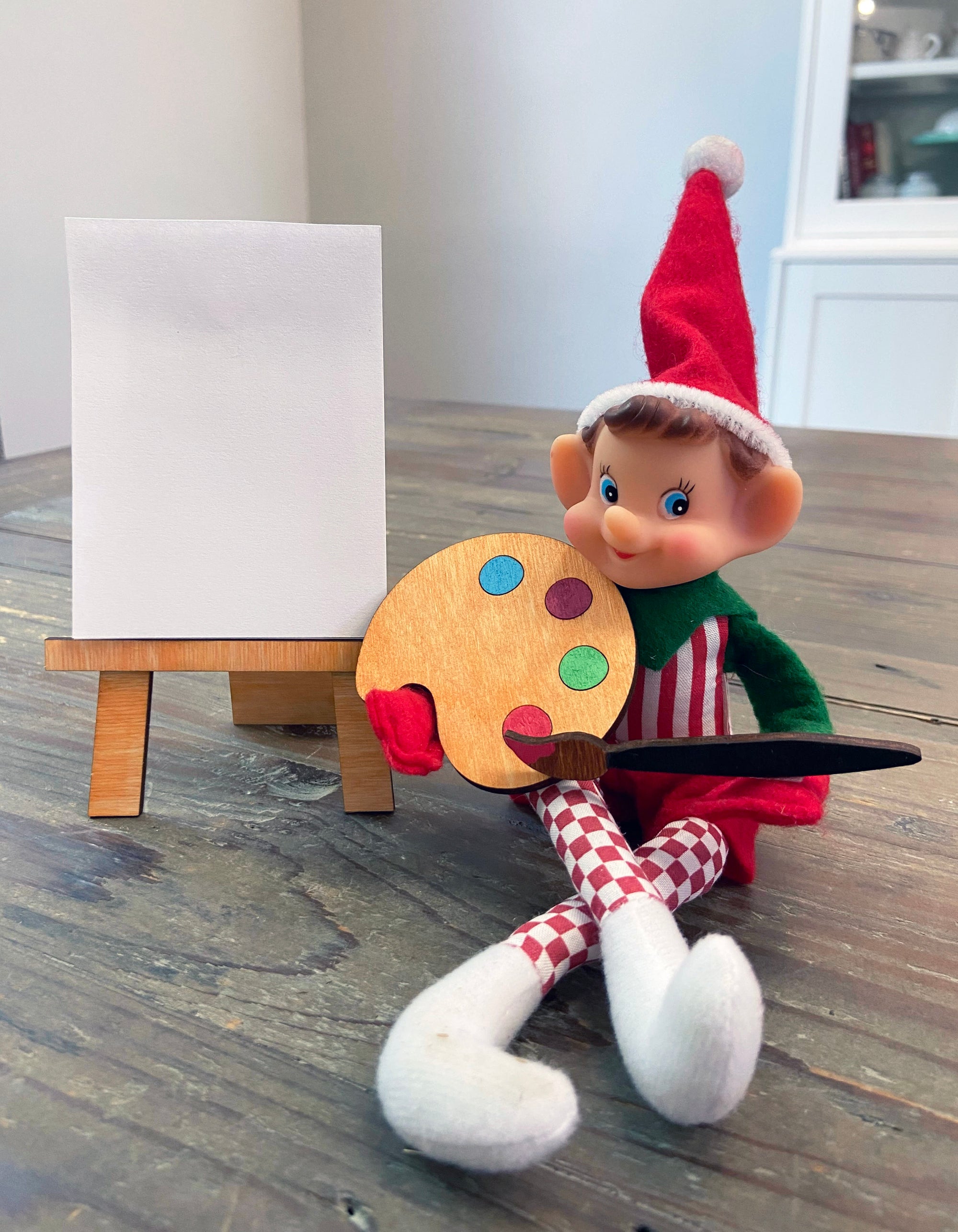 The Rustic Rhode DIY Elf Prop Kit features a festive red and white elf doll at a table with a paint palette, brush, and blank canvas on a mini wooden easel—ideal for holiday displays or pairing with handcrafted elf decor.