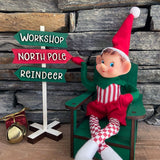 The Rustic Rhode's DIY Elf Prop Kit features a smiling elf in red and green on a green chair by a signpost labeled Workshop, North Pole, and Reindeer, plus charming handcrafted props and a gold bell for festive holiday displays.