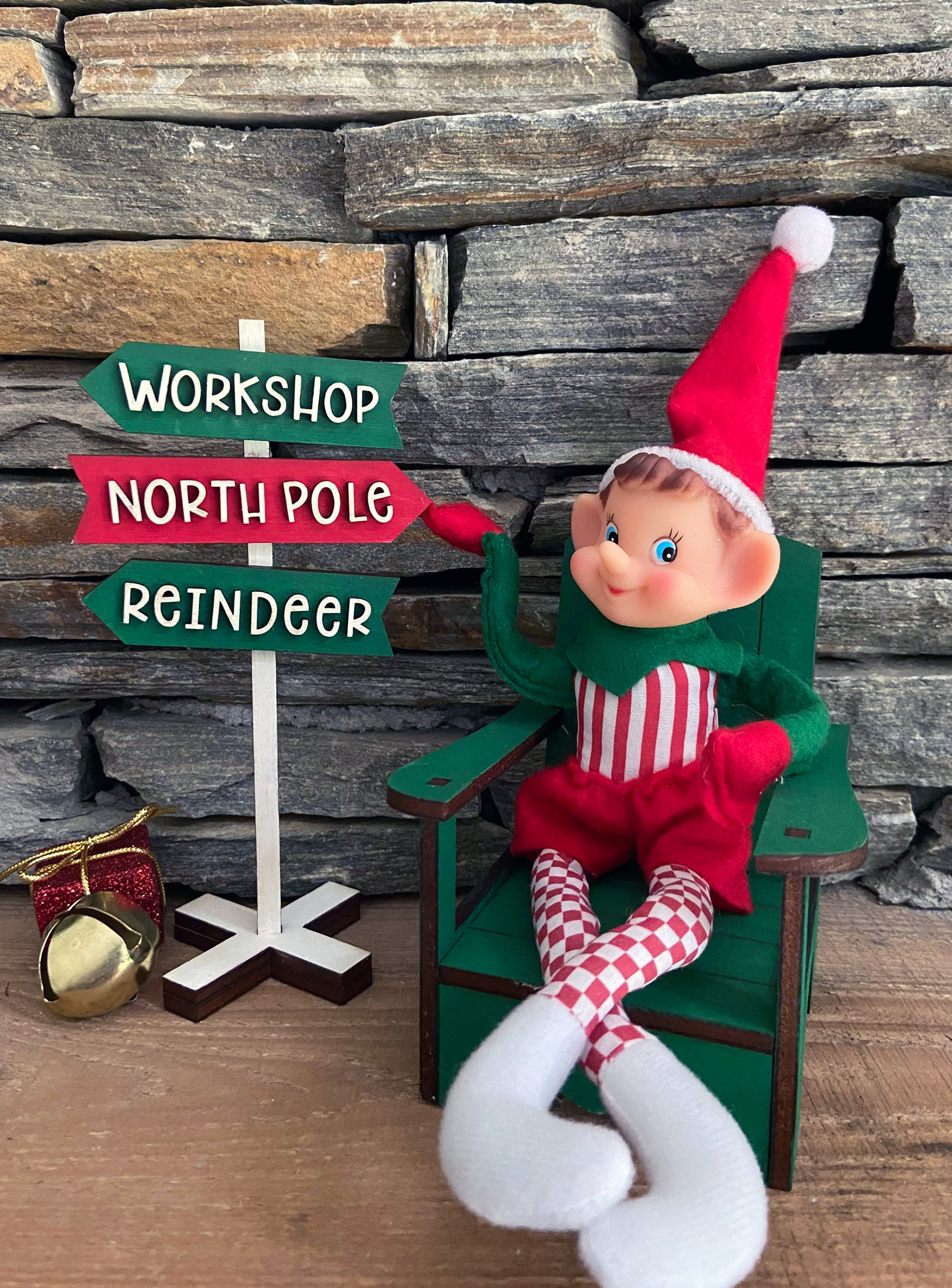 The Rustic Rhode's DIY Elf Prop Kit features a smiling elf in red and green on a green chair by a signpost labeled Workshop, North Pole, and Reindeer, plus charming handcrafted props and a gold bell for festive holiday displays.