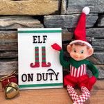 A plush elf doll from The Rustic Rhode's DIY Elf Prop Kit sits beside an "Elf on Duty" sign with striped elf legs, ideal for holiday displays, against a stone backdrop with a gold present and jingle bell.