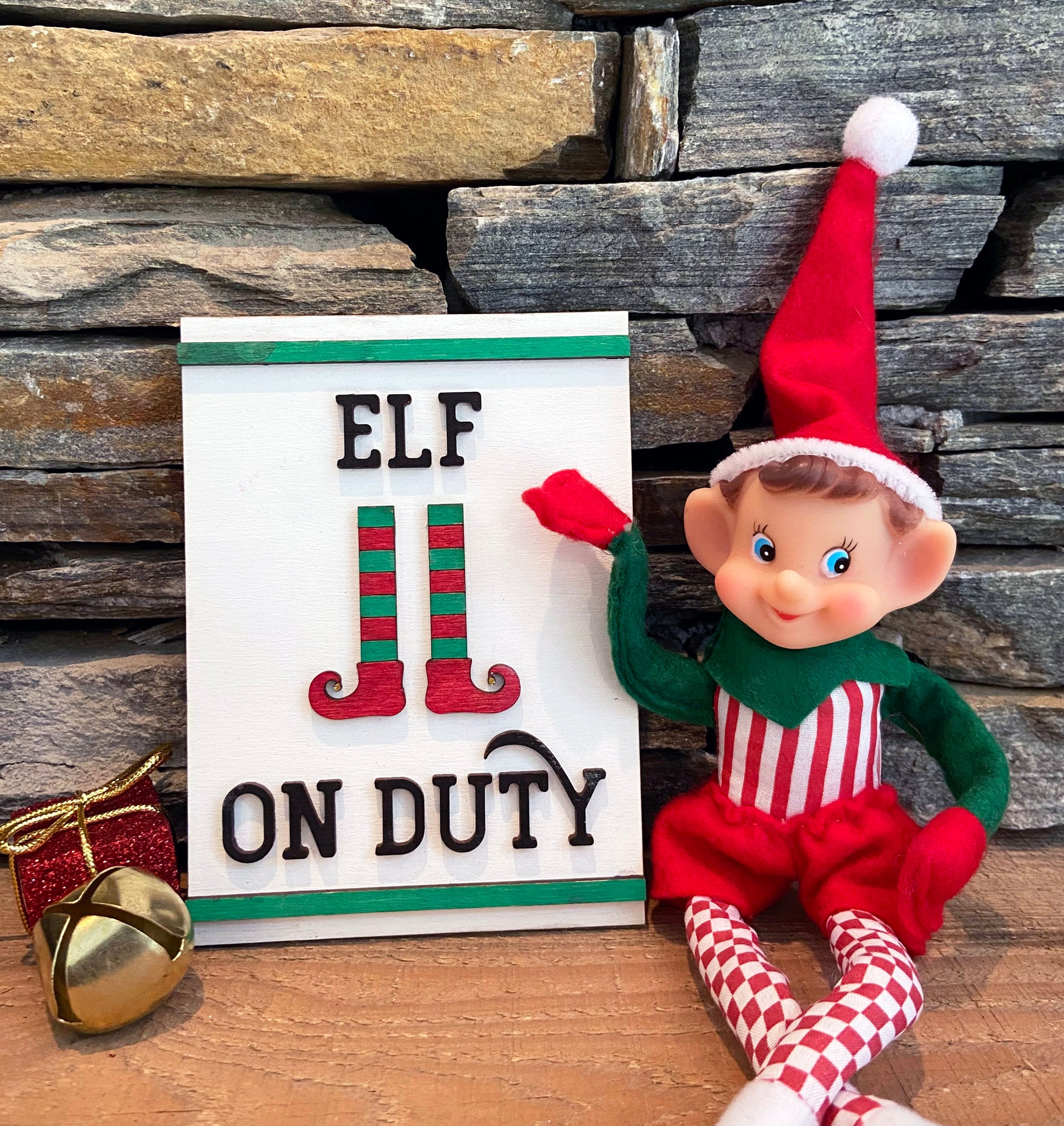 A plush elf doll from The Rustic Rhode's DIY Elf Prop Kit sits beside an "Elf on Duty" sign with striped elf legs, ideal for holiday displays, against a stone backdrop with a gold present and jingle bell.