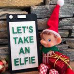 The Rustic Rhode's DIY Elf Prop Kit features a festive elf doll in a red hat with a "LETS TAKE AN ELFIE" sign, wrapped gifts, and stone wall backdrop—ideal for holiday displays or pairing with handcrafted elf decor.