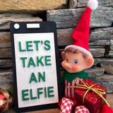 The Rustic Rhode's DIY Elf Prop Kit features a festive elf doll in a red hat with a "LETS TAKE AN ELFIE" sign, wrapped gifts, and stone wall backdrop—ideal for holiday displays or pairing with handcrafted elf decor.