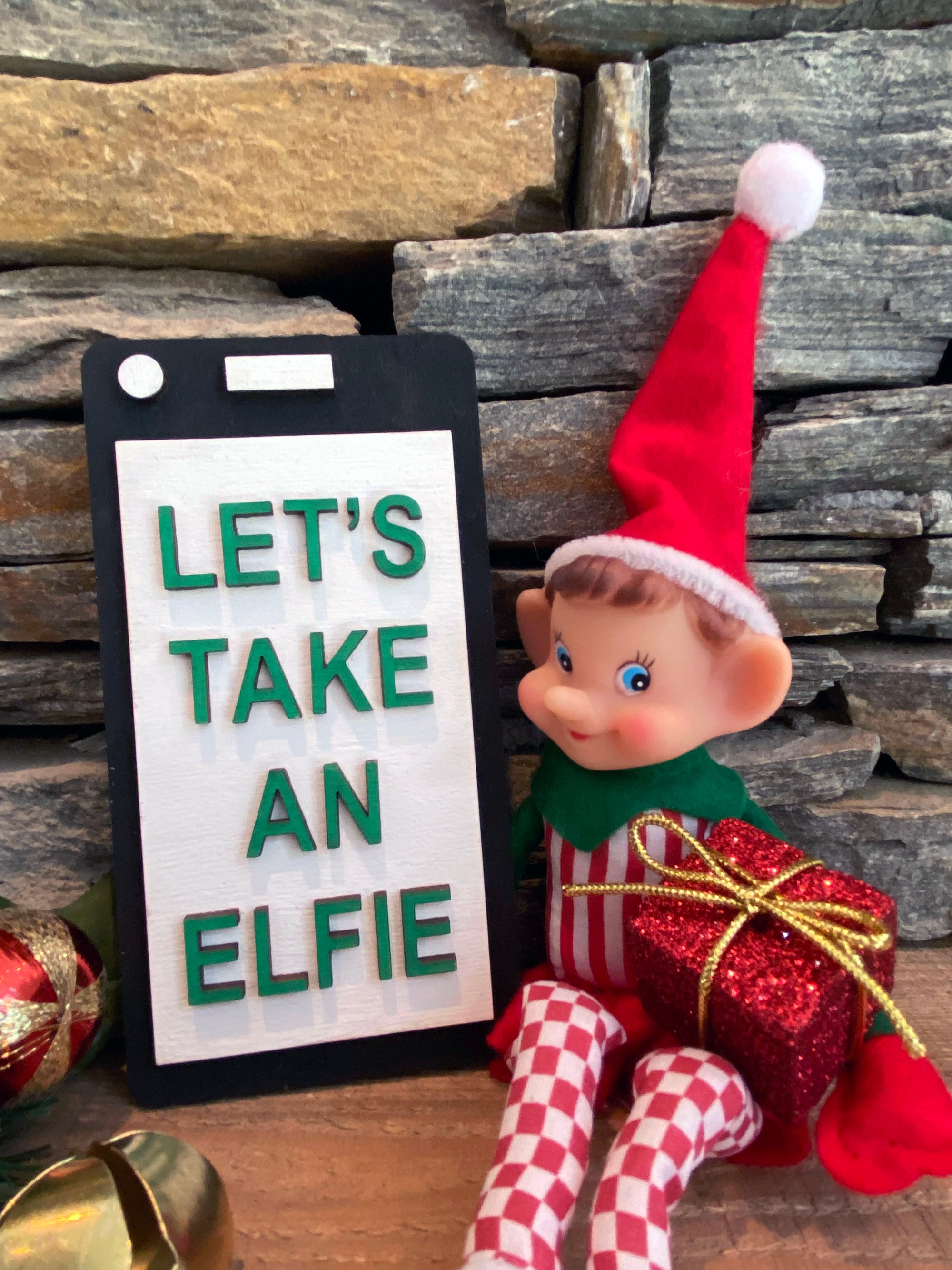 The Rustic Rhode's DIY Elf Prop Kit features a festive elf doll in a red hat with a "LETS TAKE AN ELFIE" sign, wrapped gifts, and stone wall backdrop—ideal for holiday displays or pairing with handcrafted elf decor.