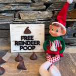 The Rustic Rhode DIY Elf Prop Kit features a smiling Christmas elf doll sitting by a "Free Reindeer Poop" sign with chocolate candy, plus handcrafted elf props and chocolates—perfect for festive holiday displays.