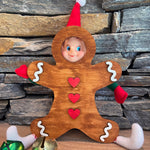 A playful elf doll made with The Rustic Rhode DIY Elf Prop Kit peeks through a wooden gingerbread man cutout, while handcrafted props and colorful jingle bells complete this festive holiday display against a stone wall.