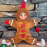 A playful elf doll made with The Rustic Rhode DIY Elf Prop Kit peeks through a wooden gingerbread man cutout, while handcrafted props and colorful jingle bells complete this festive holiday display against a stone wall.