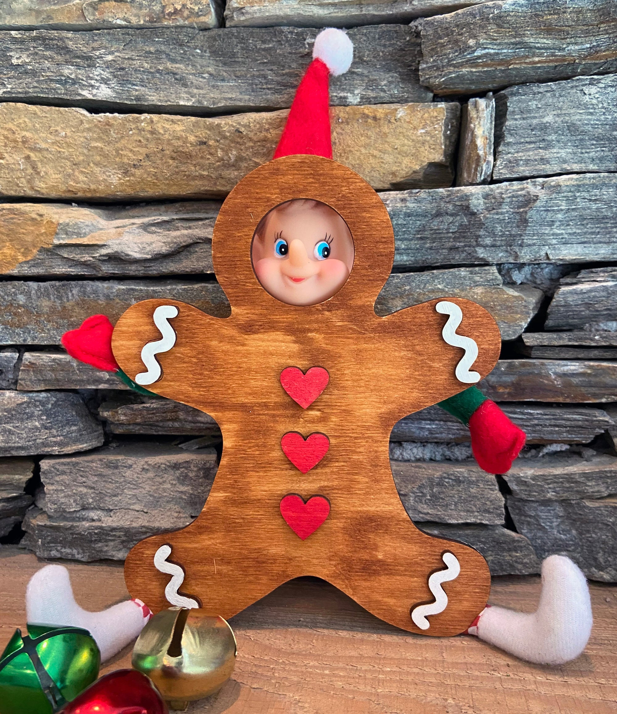 A playful elf doll made with The Rustic Rhode DIY Elf Prop Kit peeks through a wooden gingerbread man cutout, while handcrafted props and colorful jingle bells complete this festive holiday display against a stone wall.