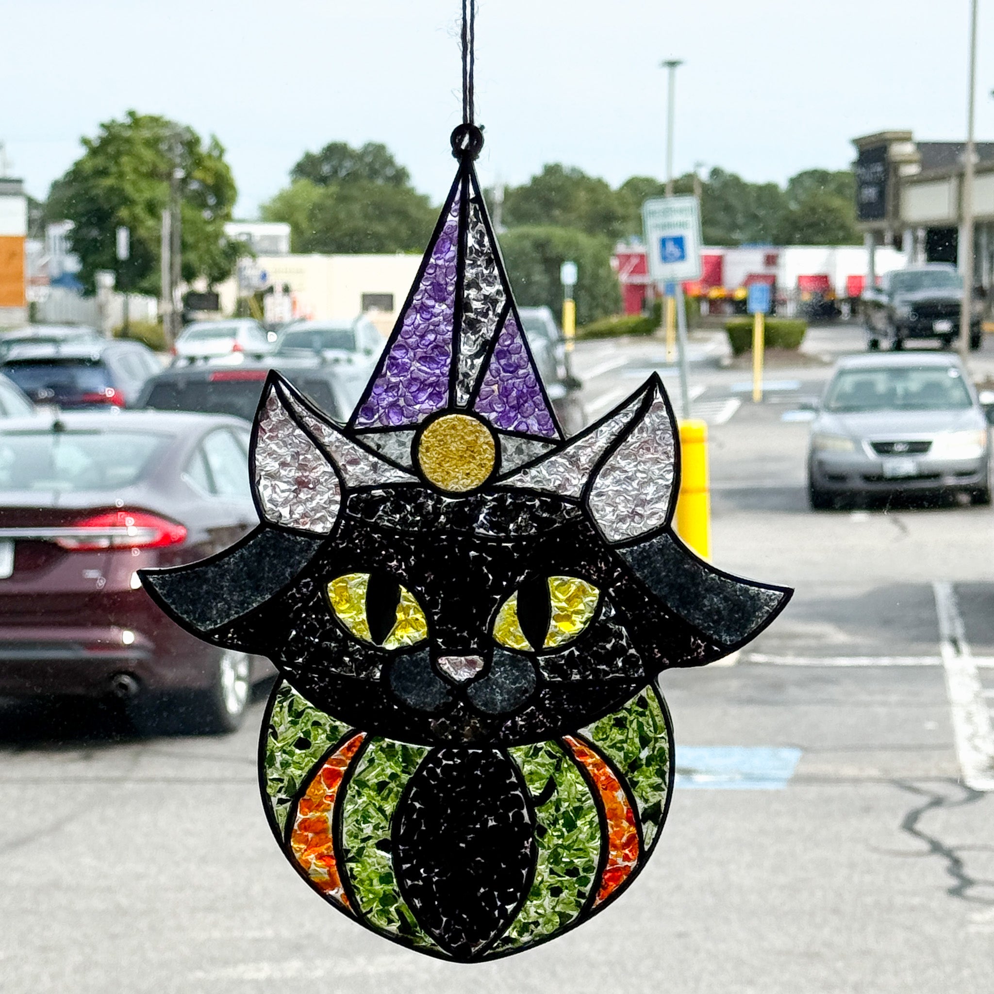 The Rustic Rhode's Autumn Magic Suncatchers Workshop features a black cat suncatcher with yellow eyes and a purple wizard hat—perfect for Halloween crafting fun, seen hanging in a window with parked cars outside.