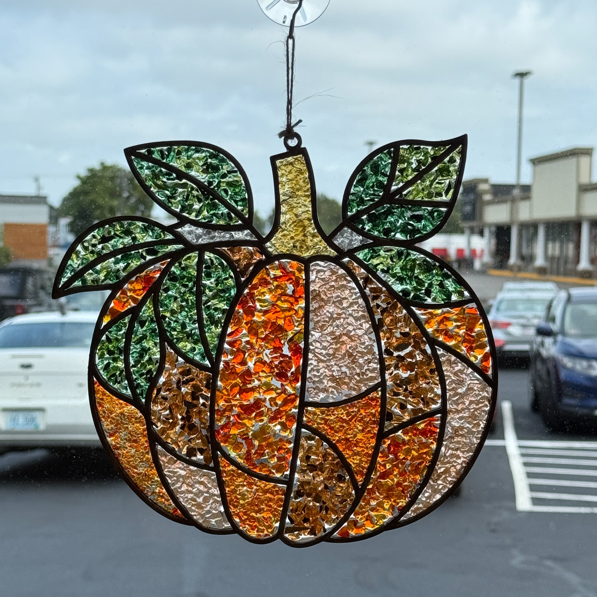 A pumpkin-shaped suncatcher with green leaves from The Rustic Rhode’s Autumn Magic Suncatchers Workshop hangs on a window, with cars and buildings visible outside under a cloudy sky.