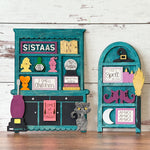 Two teal and black wooden shelves from The Rustic Rhode display colorful Halloween decor—witches, hats, potion bottles, and SISTAAS signs—inspiring creations at a Hocus Pocus Craftus Workshop.