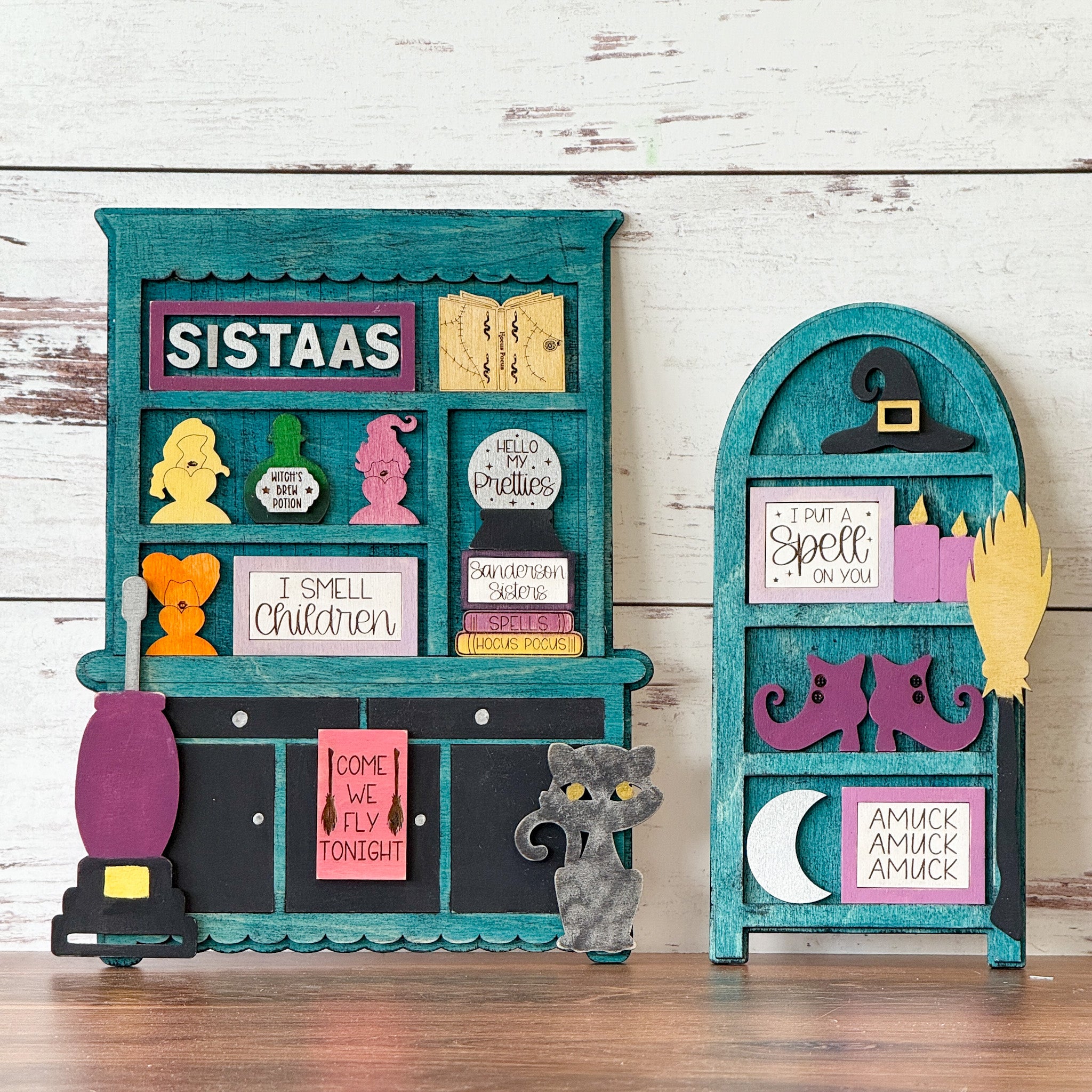 Two teal and black wooden shelves from The Rustic Rhode display colorful Halloween decor—witches, hats, potion bottles, and SISTAAS signs—inspiring creations at a Hocus Pocus Craftus Workshop.