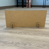 A rectangular plain brown cardboard from The Rustic Rhode’s DIY Grinch Believe kit stands upright on a light wood floor with two slotted stands, ideal as a base for holiday décor. Shelves and part of a tiled wall appear in the background.