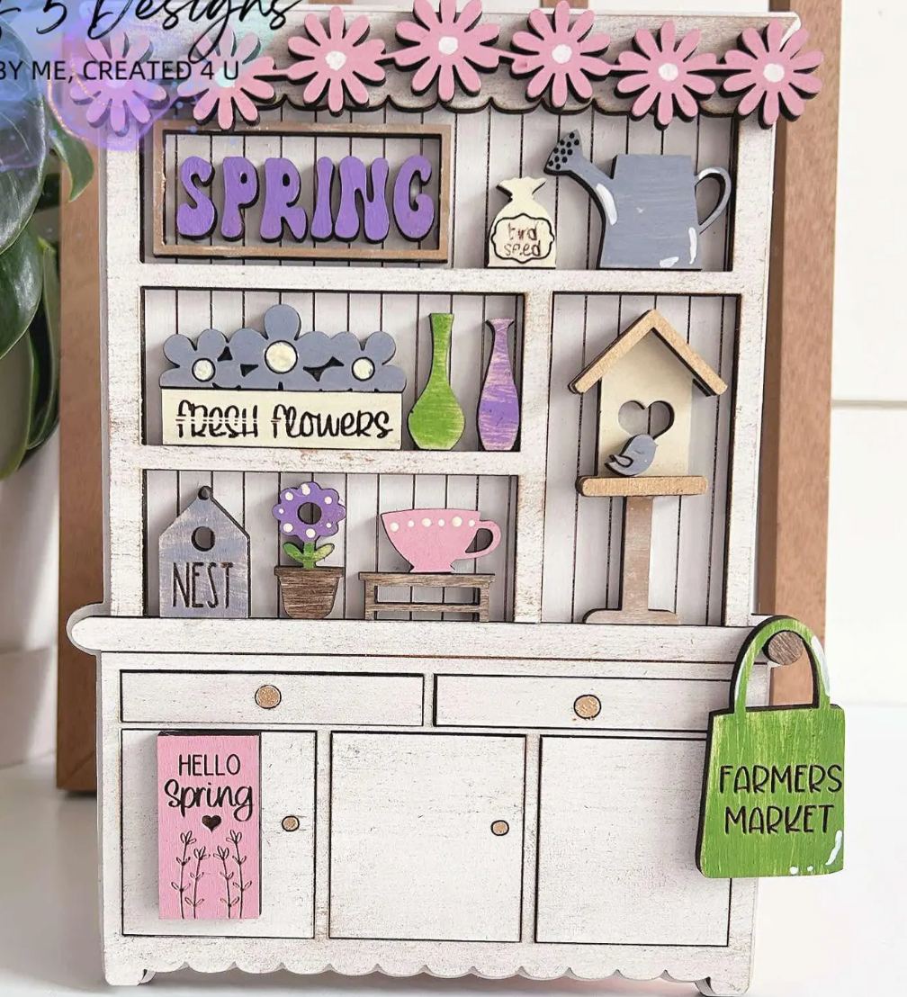 The Rustic Rhode P. Fallon Group is a miniature wooden hutch adorned with spring florals, a watering can, birdhouse, and cheerful signs—perfect for farmhouse-inspired décor or your seasonal project menu.
