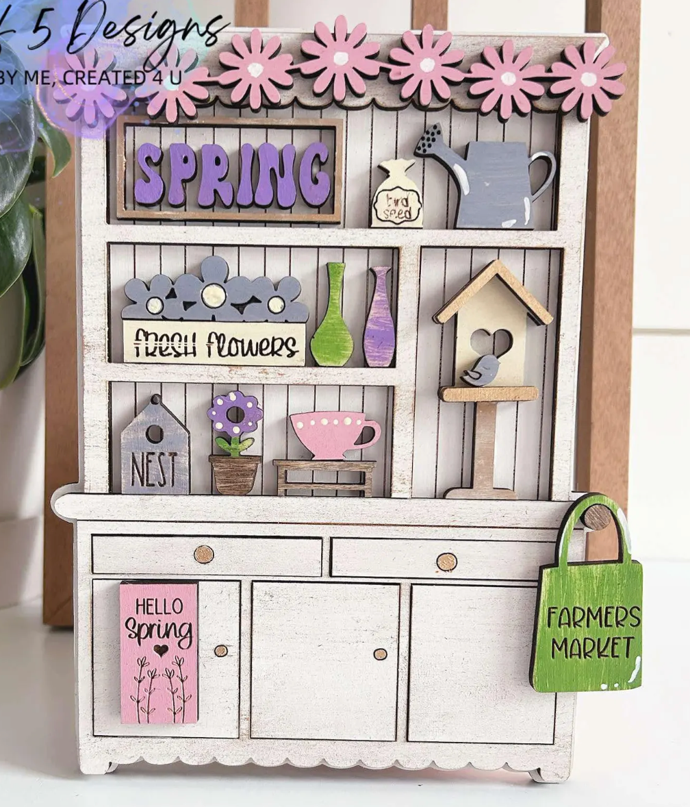 The Rustic Rhode P. Fallon Group is a miniature wooden hutch adorned with spring florals, a watering can, birdhouse, and cheerful signs—perfect for farmhouse-inspired décor or your seasonal project menu.