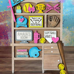 A wooden shelf featuring The Rustic Rhode’s Warwick Softball Fundraiser-10/2/25 door hangers, colorful softball-themed signs, trophies, a glove and bat with softballs, plus phrases like “Home Run” and “There’s no crying in softball.”.