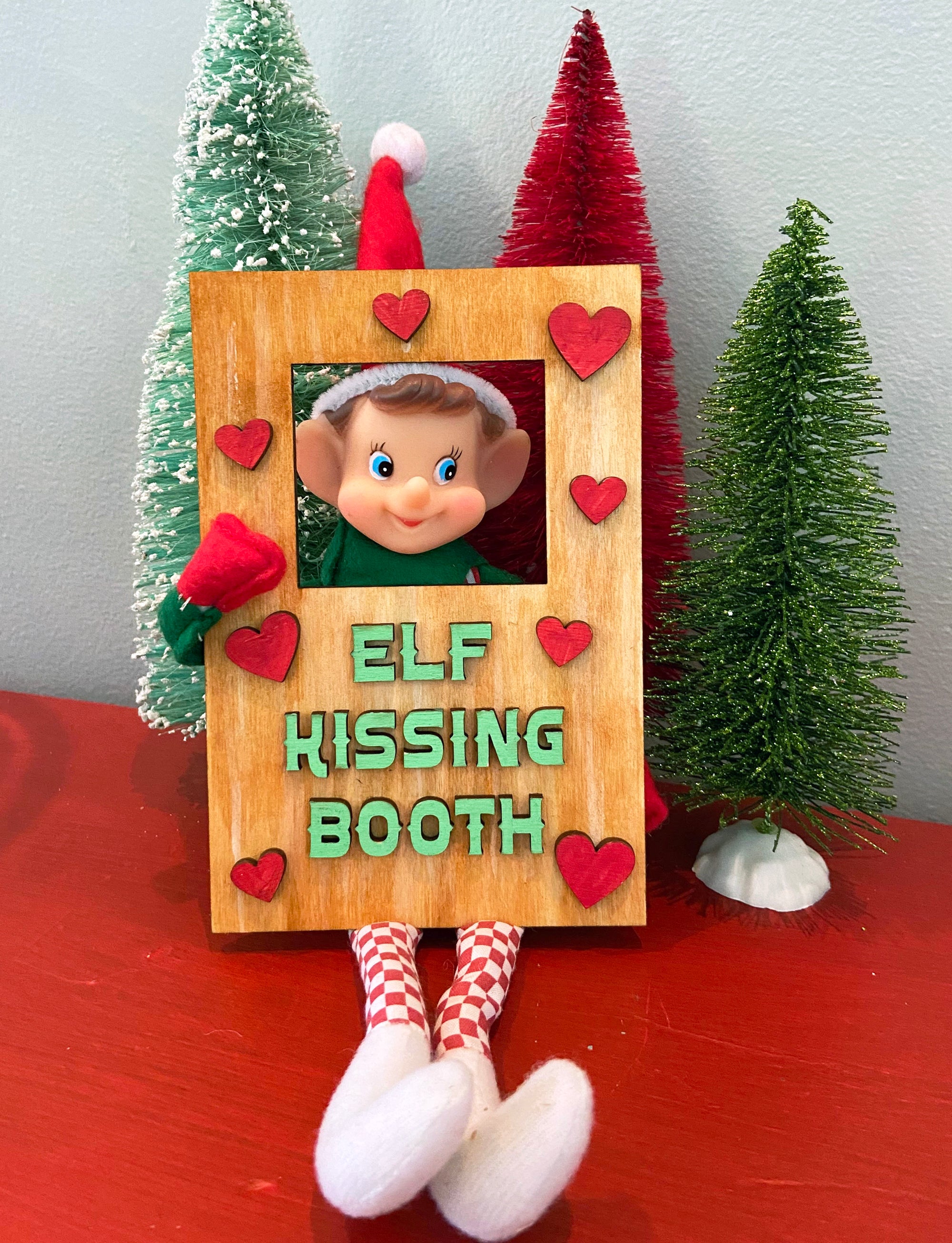 Create a charming holiday display with The Rustic Rhode DIY Elf Prop Kit—featuring a festive elf doll in a wooden Elf Kissing Booth with red heart accents and three mini Christmas trees, perfect for your Christmas décor.