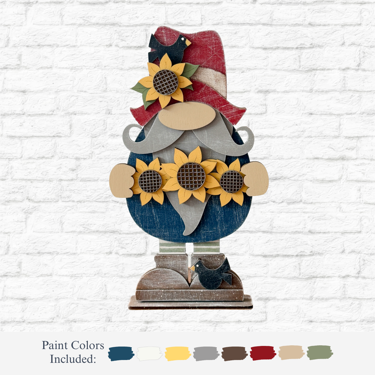 The Rustic Rhode’s DIY Standing Sunflower Gnome features a blue body, red and white striped hat, sunflowers, and birds at its feet. Laser cut wood, shown against a white brick wall with six paint colors included.