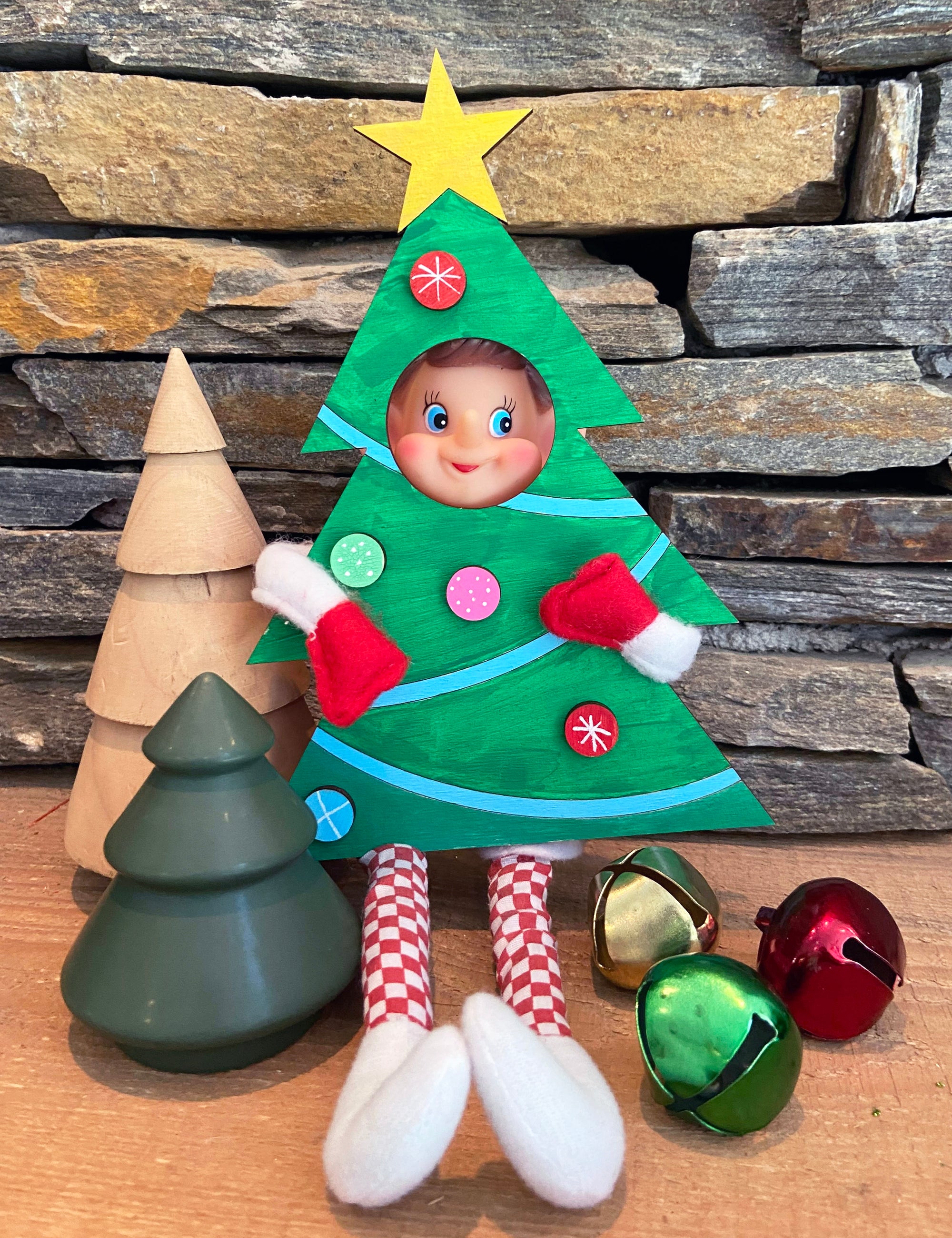 The Rustic Rhode’s DIY Elf Prop Kit features a doll dressed as a Christmas tree beside stacked stones, wooden and green tree decorations, and jingle bells—perfect for creative holiday displays or pairing with handcrafted elf props.