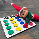 A whimsical elf doll with a red hat and striped pants lies on a wooden Elf Twister board, featuring colorful circles, handcrafted by The Rustic Rhode—perfect for holiday displays or pairing with the DIY Elf Prop Kit.