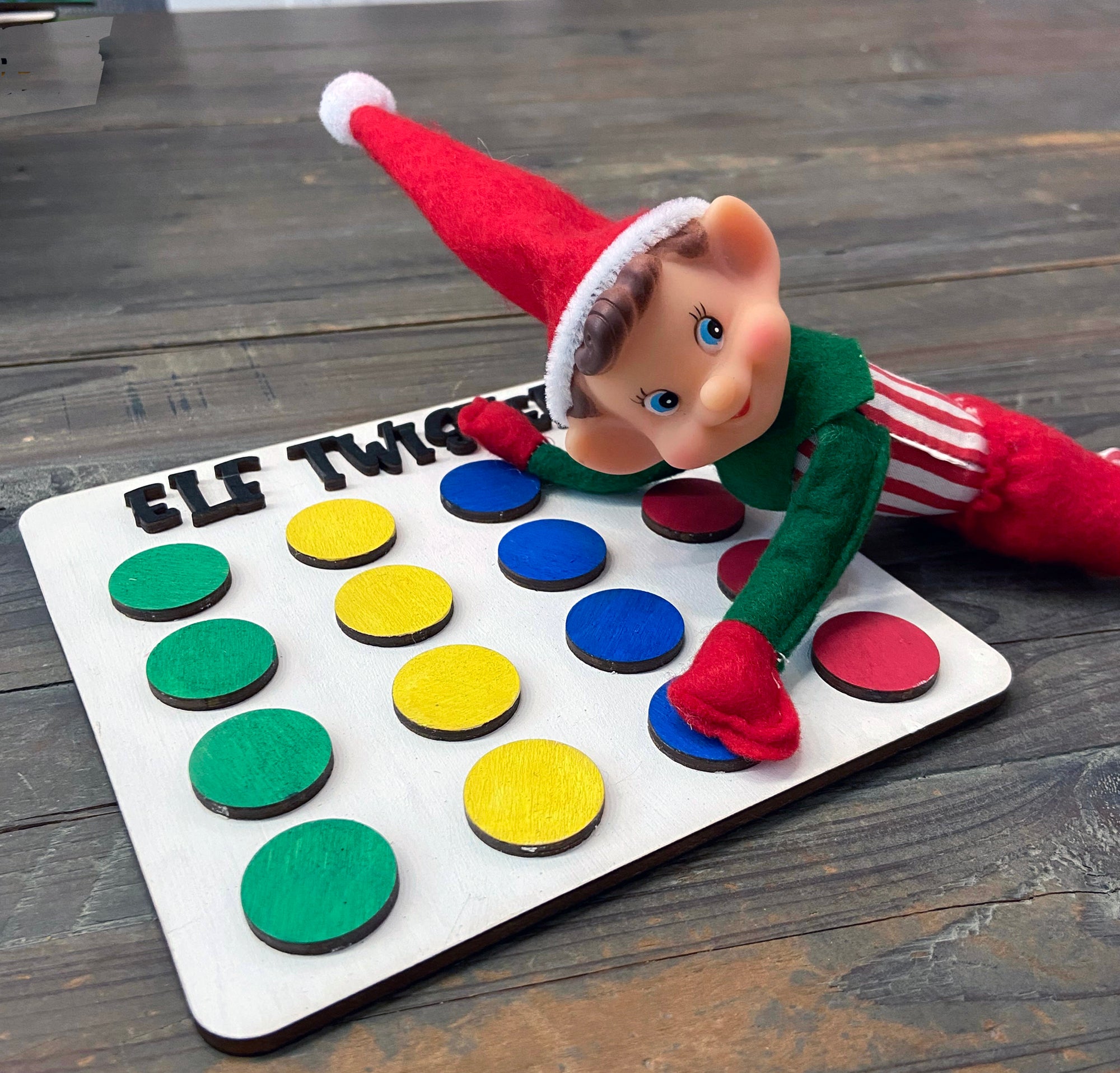 A whimsical elf doll with a red hat and striped pants lies on a wooden Elf Twister board, featuring colorful circles, handcrafted by The Rustic Rhode—perfect for holiday displays or pairing with the DIY Elf Prop Kit.