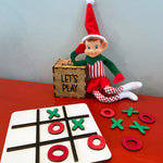 The Rustic Rhode's DIY Elf Prop Kit features an elf doll in red and green, a wooden "LETS PLAY" box, and a tic-tac-toe board with red O and green X pieces—ideal for holiday displays or festive crafts.