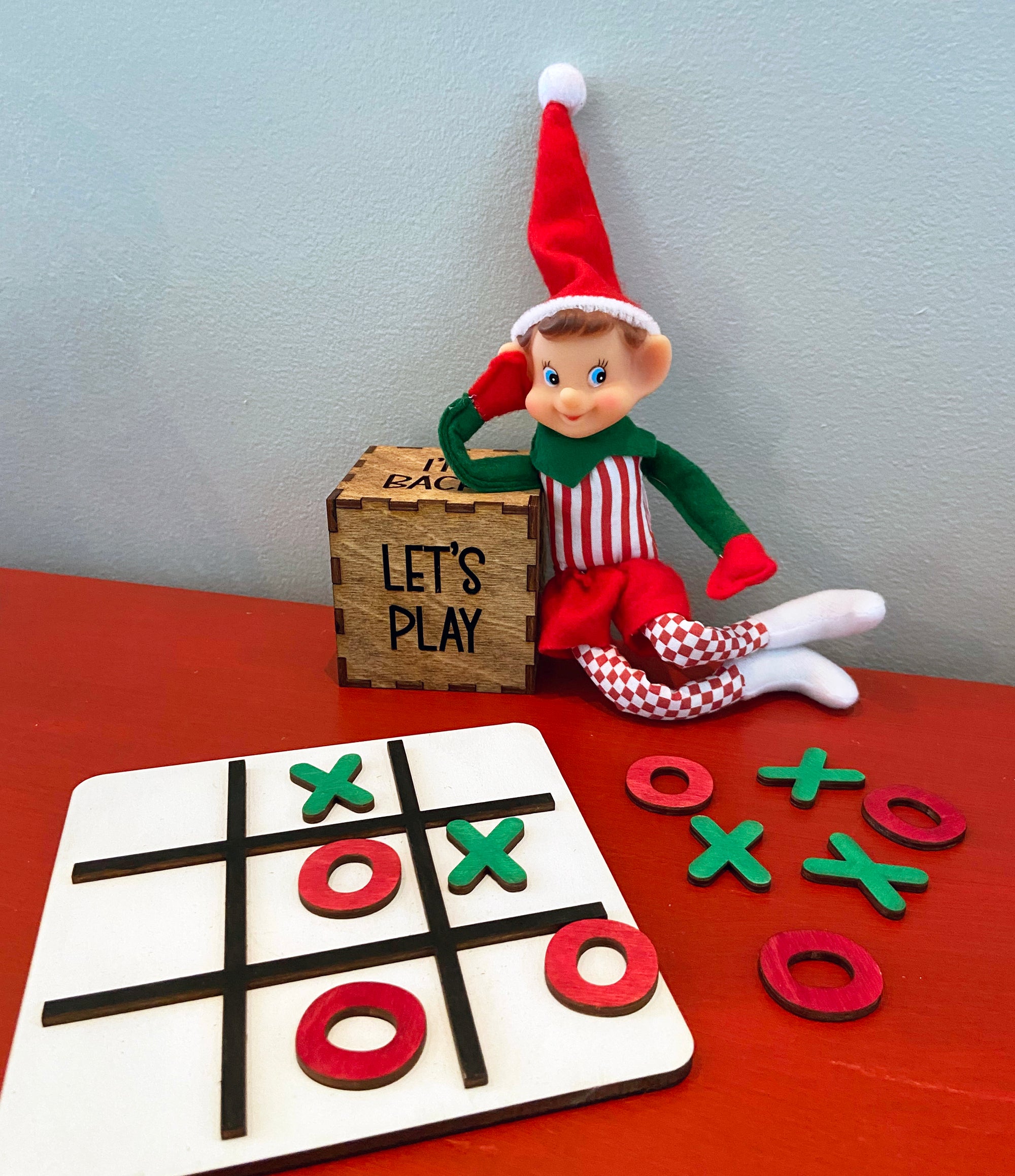 The Rustic Rhode's DIY Elf Prop Kit features an elf doll in red and green, a wooden "LETS PLAY" box, and a tic-tac-toe board with red O and green X pieces—ideal for holiday displays or festive crafts.